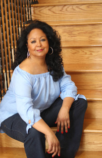 Michele Maddox in a light blue peasant blouse and black pants sits casually, with her left knee up and left arm resting on her knee. She is smiling at the camera.